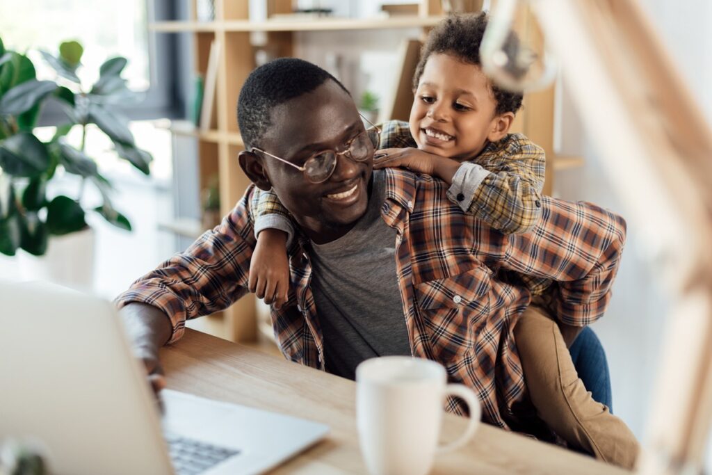 A dad and son playing while the dad works remotely from his home. This promotes work life balance as a part of a total rewards strategy.